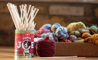 A can with a dog graphic holds knitting needles, with a ball of burgundy yarn and a box of colorful skeins of yarn in the background on a wooden surface.