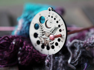 A close-up of the 🌙 Night Sky & Forest Floor Knitting Needle Gauge 🌙—a circular cherry wood tool with numbered and plant engravings—rests on purple and blue yarn, knitting needles, and features subtle chanterelle mushroom etchings.