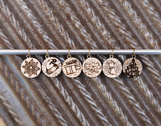 The Maple Wood Yuletide Knitting Stitch Markers Set of 6 features engraved holiday designs—snowflake, candy cane, gift box, string lights, reindeer, and Merry Xmas tree—displayed in a row on a metal rod against a textured background.