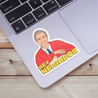 A laptop displays the "Hey Neighbor | Mr. Rogers Sticker," featuring a smiling Mr. Rogers in a red cardigan waving with "HEY NEIGHBOR" in bold yellow letters, resting on a wooden surface.