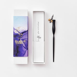 The Flourish Calligraphy Pen is a beginner set with an oblique pen, black barrel, brass nib, and comes in a white box with purple and blue watercolor art plus a gold-embossed logo, all displayed on a white surface.