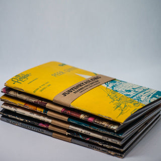 A stack of colorful booklets rests on a white surface, with the top featuring a bright yellow cover and brown paper band—evoking the design of the Vintage Vinyl Pocket Notebook.