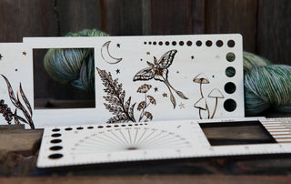 The Luna Moth “Almost Everything” Ruler is a wooden fiber arts tool with engraved luna moth, moon, stars, plants, and mushrooms designs. It features needle sizing holes and a ruler—shown in front of green and gray yarn.