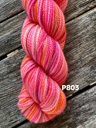 A skein from the KPM and KPPPM Variegated Color Collection is shown on a rustic wooden surface in pink, red, orange, and yellow shades. Perfect for hand knitting, the code P803 appears in white on the right.