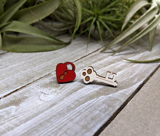 The Key and Heart Lock Earrings, made from sustainable wood, feature a red heart with a keyhole and a white key, displayed on a wooden surface with green plants in the background—both charming and eco-friendly.