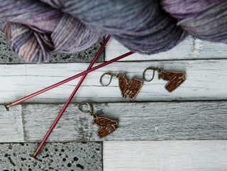 Three Conspiracy of Ravens Knitting Progress Keepers made of maple wood rest on a white and gray wooden surface beside two red knitting needles and a skein of purple and gray yarn, creating charming knitting accessories.