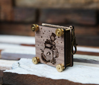 The Miniature Flower Press is a small decorative walnut wood piece with engraved art of a jar, plants, and stars. It stands upright on a rustic wooden surface and is secured with brass corner screws.