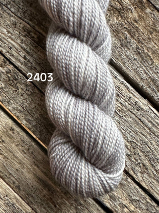 A skein of Koigu KPM & KPPPM Fingering Weight Yarn in light gray (color 2403) rests on a rustic wooden surface with visible texture and grain.