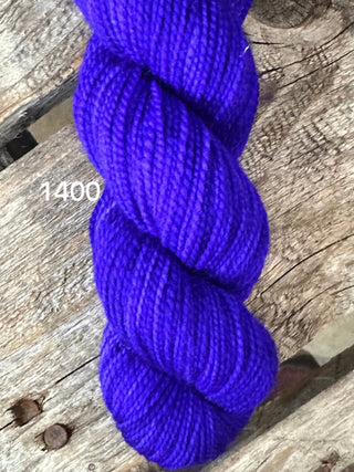 A close-up of a skein of vibrant purple Koigu KPM & KPPPM Fingering Weight Yarn labeled “1400,” resting on a rustic wooden surface.