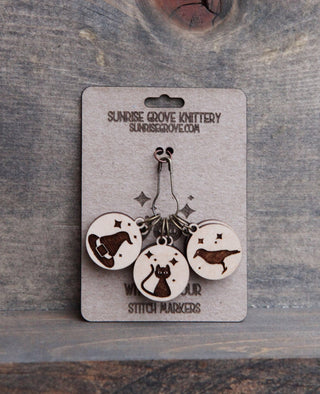 The Witching Hour Stitch Markers from Sunrise Grove Knittery include three maple wood markers engraved with a yarn ball, cat, and seal, shown as witch-themed knitting accessories on a rustic wood background.