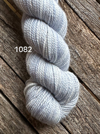A skein of Koigu KPM & KPPPM Fingering Weight Yarn in light gray-blue (color 1082) rests on a rustic wooden surface—ideal for your next knitting or crochet project.