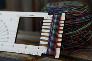 A close-up shows multicolored yarn wrapped around the Butterfly "Almost Everything" Ruler, with a matching yarn ball in the background. The ruler features inch marks and slots, serving as a convenient knitting gauge for measuring yarn thickness.