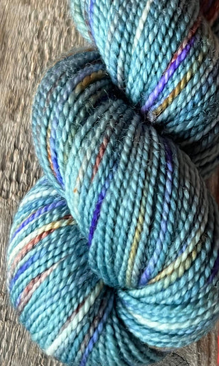 A close-up of KPM and KPPPM Variegated Color Collection Skeins in blue with multicolored speckles—purple, yellow, and orange—on a textured gray surface, ideal for hand knitting soft merino wool projects.