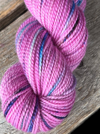 A close-up of a KPM and KPPPM Variegated Color Collection Skein in pink with blue, purple, and gray streaks rests on rustic wood—ideal for hand knitting projects.