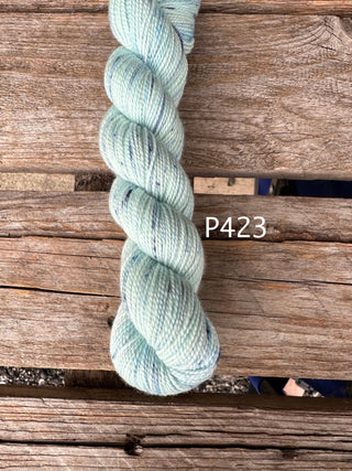 A twisted skein of pale mint green Bridgerton Collection KPPPM yarn with subtle flecks, perfect for hand knitting. Resting on weathered wooden boards, the label P423 is displayed next to the merino wool yarn.