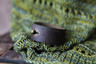 The Bee and Moon Leather Ruler Cuff, a wide dark brown reclaimed leather bracelet with a brass stud closure, rests on textured green knitted fabric.