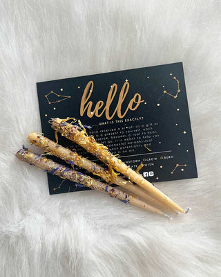 Three Natural beeswax ritual candles with dried flowers and gold flakes rest on white faux fur, in front of a dark card reading hello in gold with constellation art—an ideal addition to spiritual practices.