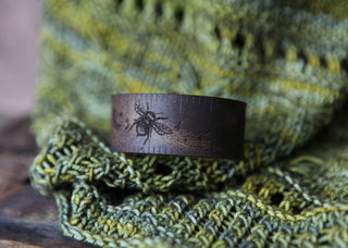 The Bee and Moon Leather Ruler Cuff, made from reclaimed leather, features an engraved bee design and is shown resting on textured green knitting for a unique, eco-friendly look.