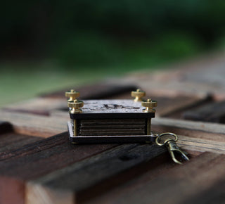 The Miniature Flower Press, a small vintage-style walnut wood keychain with metallic corners and screw details, rests on a wooden surface against a blurred green background.