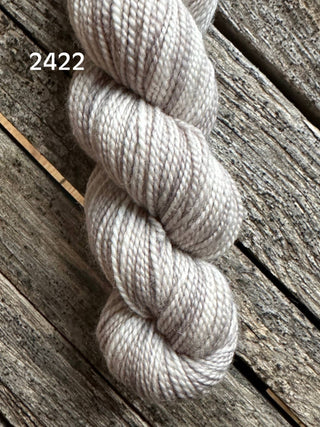 A skein of light gray, slightly shiny Koigu KPM & KPPPM Fingering Weight Yarn (color 2422) is lying on a rough wooden surface with visible grain and texture.