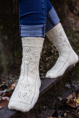 Feet-only view of a woman balancing on a piece of wood, jeans rolled up to show off a pair of knitted socks. The socks are a pale yellow yarn, hand dyed by Cat Tails yarn, with a delicate floral motif by Liz Harris