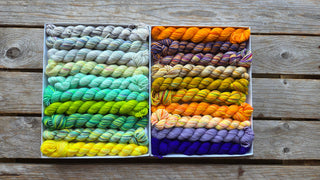 Two white boxes of Spooky Season Koigu Pencil Boxes sit on wood, each filled with colorful mini skeins of Merino Wool—cool greys, greens, and yellows on the left; warm orange, purple, and gold hues on the right.