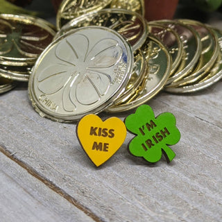 A yellow heart “Kiss Me” pin and a green “I’m Irish” shamrock pin rest beside gold coins with four-leaf clovers—festive accents to pair with Kiss Me I’m Irish Earrings St. Patrick's Day, handmade in the USA.