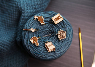 A ball of blue yarn rests on a dark surface, accompanied by five Princess Bride Knitting Stitch Markers in the shapes of a cat, sword, mask, book, and crown. Nearby are a crochet hook and knitted blue fabric—an ideal setup for anyone wanting to enrich their collection with these enchanting knitting accessories.