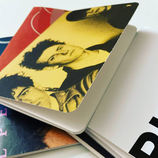 A close-up of overlapping books or magazines, with one cover showing a yellow-tinted, vintage-style photo of two people—similar to a Vintage Vinyl Pocket Notebook. The edges curl slightly, and partial text appears on the white cover beneath.