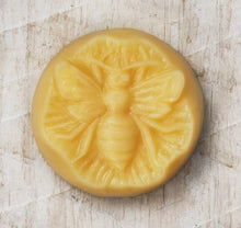 The Little Bee Lotion Bar is a round beeswax moisturizer featuring an embossed bee design, displayed on rustic light wood, and delivers deep hydration for your skin.