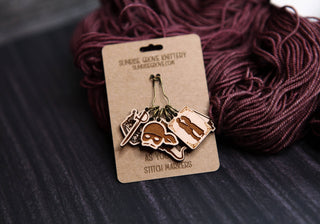 Showcased next to purple yarn, the Princess Bride Knitting Stitch Markers set includes five engraved maple wood markers on a card. Each marker displays a unique knitting-themed design, making them both charming and practical accessories for knitters.