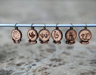 The 🔮 Crystal Ball Stitch Marker Set 🔮 features six cherry wood markers, each engraved with a moon, crystals, eye, potion bottle, bat, and mushrooms. These witchy knitting accessories hang from a rod against a blurred marble background.