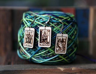 🔮 Tarot Card Stitch Markers – The High Priestess, The Empress & The Emperor rest on blue and green yarn—perfect maple wood progress keepers for knitting and crochet. Wooden surfaces and blurred objects fill the background.