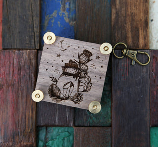 The Miniature Flower Press, a wooden keychain with brass rivets and engraved designs of a jar, cactus, crystals, and stars, sits on a colorful wooden background—perfect for preserving tiny blooms on the go.