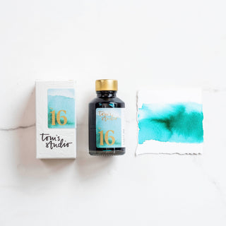 A bottle of Bottled Ink in Sunny Teal with a gold cap, its matching box, and a paper showing a teal swatch are arranged on a white surface, highlighting the vibrant Sunny Teal shade.