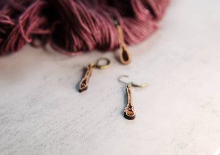 Three gold-toned 🧹 Witch’s Broom Stitch Markers 🧹 rest on a light surface beside burgundy yarn—perfect for adding charm to your knitting or crochet projects.