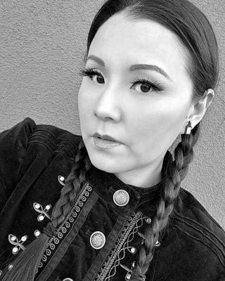 Black and white photo of a woman with long dark hair in braids, wearing a decorative geometric embroidered jacket and Wednesday Stud Earrings, looking directly at the camera against a plain wall.