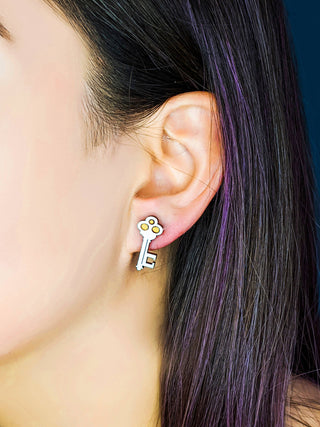 A close-up shows a person's ear with straight, dark hair and a purple streak, wearing the Key and Heart Lock hypoallergenic gold earring shaped like a key. The background is dark and blurred.