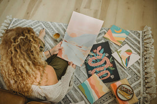 A person with curly hair sits on a patterned rug, enjoying mindful painting with the Vibrant Butterflies Meditative Art Paint by Number Kit, surrounded by colorful pottery and abstract nature scenes as creative self-care.