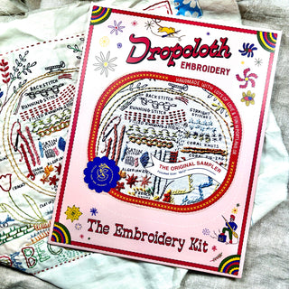 The Original Embroidery Sampler Kit box, featuring Rebecca Ringquist's stitch designs with rainbows and flowers, sits atop a partially completed embroidery project.
