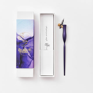 The Flourish Calligraphy Pen, featuring a purple body and metal nib ideal for beginners, sits beside an open white box with a small tree and cursive text. The box lid displays purple and blue artwork nearby.
