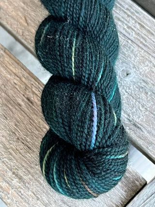 A close-up of a dark teal skein from the KPM and KPPPM Variegated Color Collection, featuring subtle multicolored streaks, ideal for hand knitting, resting on weathered wood.