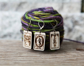 Three Tarot Card Stitch Markers—The World, The Lovers & The Hanged Man—are displayed in front of colorful yarn on a wooden surface, alongside crochet markers, with a blurred stone backdrop.