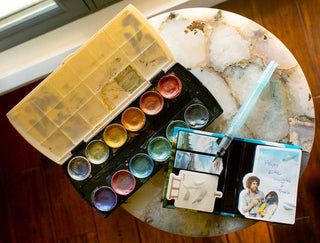 An open watercolor paint set, a notepad with an artist image and "Happy Little Thoughts," and the Bob Ross Sticky Notes booklet sit on a round, marbled table near a window.