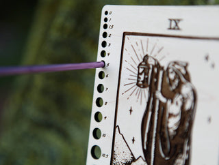A purple incense stick burns through hole 5 on ✨ The Hermit ✨ Tarot Knitting Needle Gauge & Ruler, which features a bearded figure with a lantern and numbered holes from 0 to 10 along the edge, reminiscent of a tarot card.