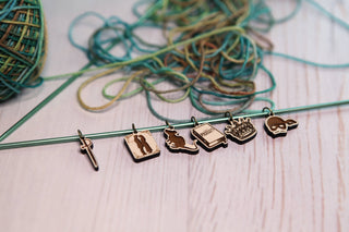 The Princess Bride Knitting Stitch Markers – featuring a set of six metal markers with designs such as a sword, silhouette, cat, book, flames, and mask – rest on a knitting needle, making an enchanting addition to any knitting accessories collection. Multicolored yarn is scattered in the background atop a light maple-like wooden surface.