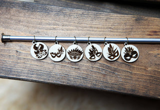 The Maple Wood Masquerade Mask Stitch Markers Set of 6 features round maple wood charms, each engraved with a unique feather or leaf Mardi Gras mask, displayed on a metal rod against wood.