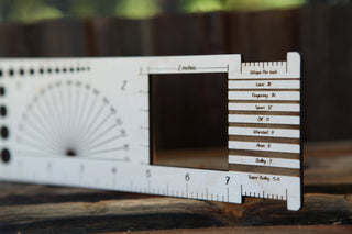 A close-up of the Luna Moth “Almost Everything” Ruler, featuring a rectangular cutout, multiple measurement markings, a semicircular protractor, and labeled gauge slots—ideal as a knitting crochet ruler—resting on a wooden surface.