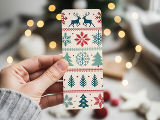 A hand holds a "Holiday Bookmarks for Knitters" printable, featuring festive red, green, and blue designs—reindeer, Christmas trees, snowflakes, and stars—while blurred Christmas lights softly glow in the background.