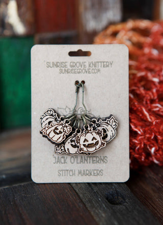 A set of cherry wood Pumpkin Jack O’Lantern Stitch Markers with carved faces is displayed on a card labeled "Pumpkin Jack O’Lantern Stitch Markers" from Sunrise Grove Knittery, resting on a rustic wooden surface.